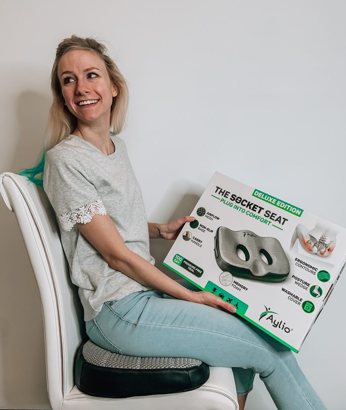 A woman sits on the Socket Seat on a white dining chair with the retail box on her lap; from the side, the gray mesh top and black non-slip base are visible.